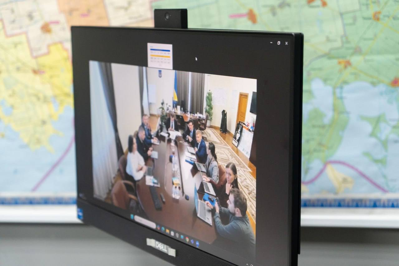 Монітор комп'ютера з відеоконференцією, де видно групу людей, які сидять навколо великого столу для нарад. На задньому плані - велика географічна карта.