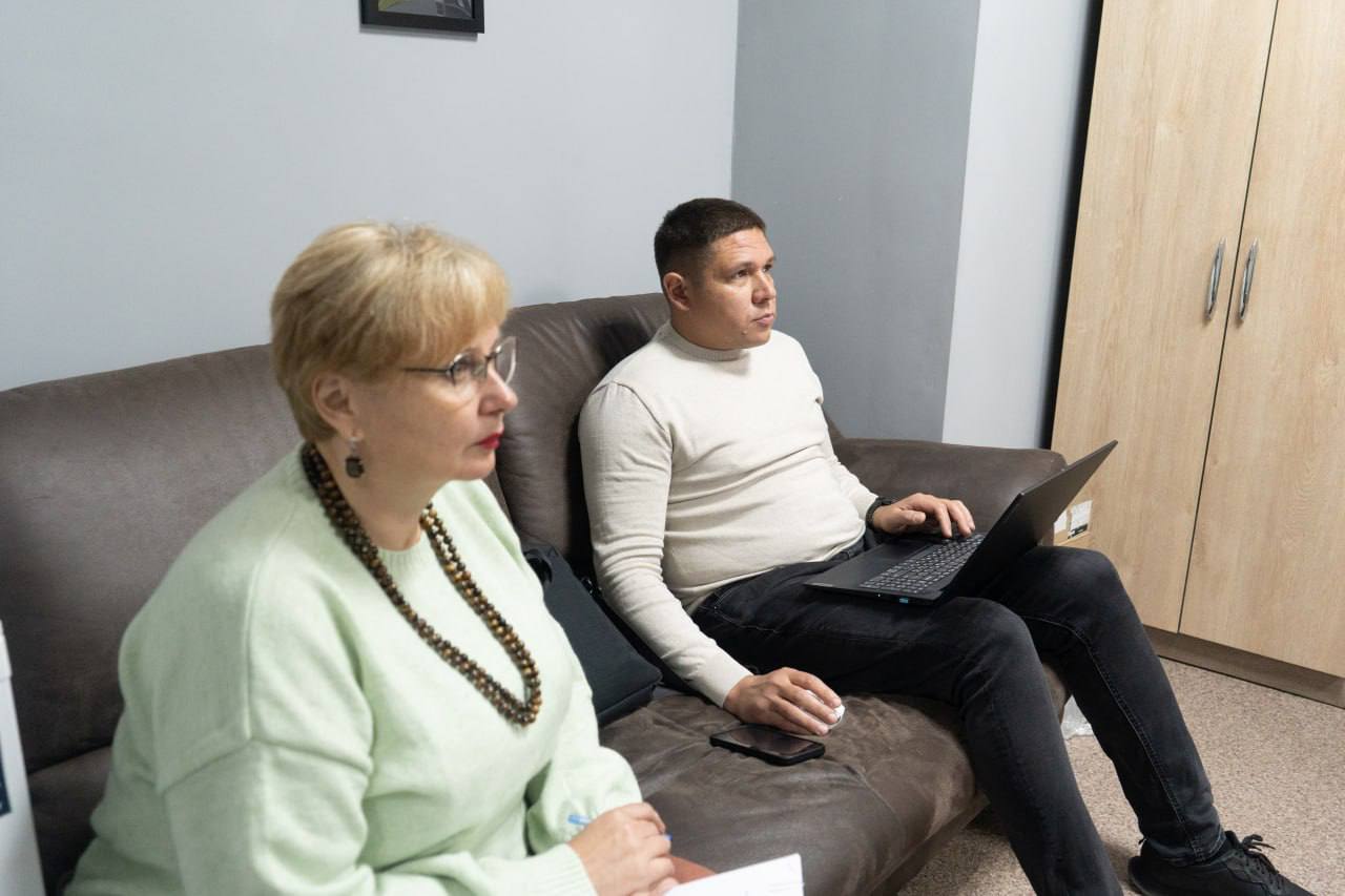 Жінка у світло-зеленому светрі та чоловік у світлому гольфі сидять на коричневому дивані. Чоловік тримає ноутбук.