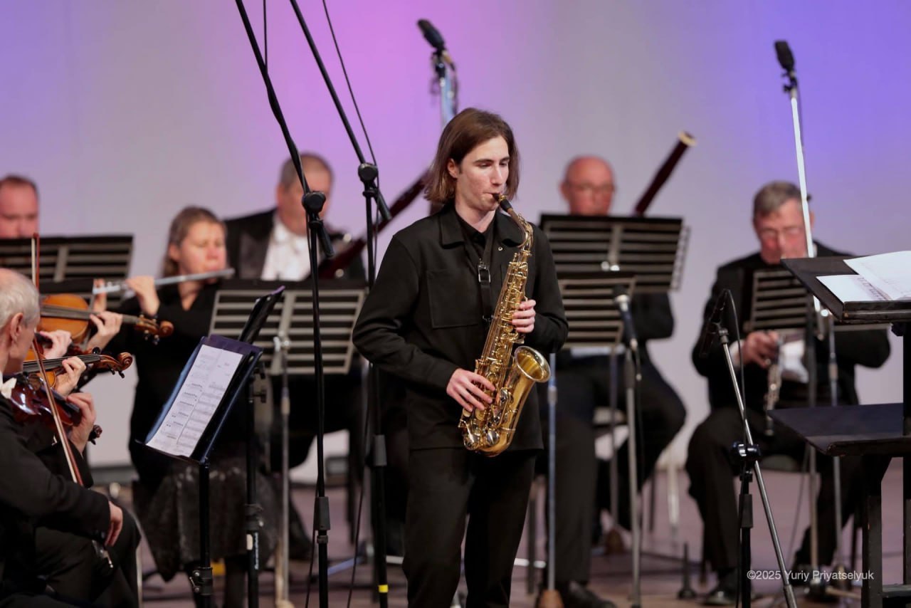 Музикант у чорному одязі грає на золотистому саксофоні на сцені у супроводі оркестру.