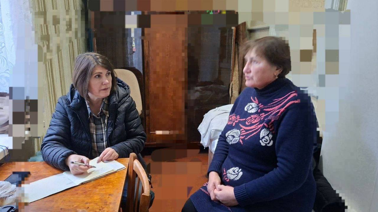 Дві жінки сидять за дерев'яним столом у приміщенні; одна, одягнена у темну стьобану куртку, записує щось у документ, інша, одягнена у синій светр з візерунком, сидить навпроти.