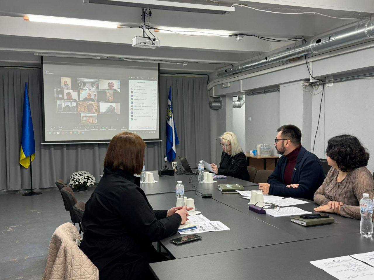 Зустріч у приміщенні: четверо людей сидять за довгим столом на фоні великого екрана з відеоконференцією. Ліворуч стоять прапор України та прапор із синьо-білим колом (можливо, обласний чи міжнародний).