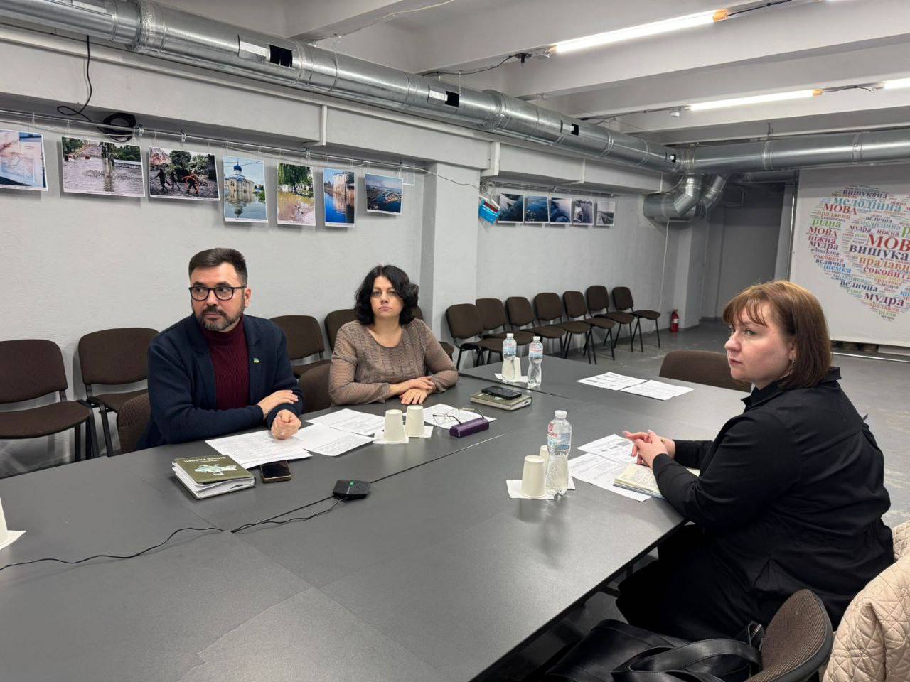 Троє людей (чоловік і дві жінки) сидять за довгим столом у приміщенні під час зустрічі чи обговорення. На стіні позаду них висять фотографії та великий плакат із текстом у формі хмари слів, що містить українські слова 