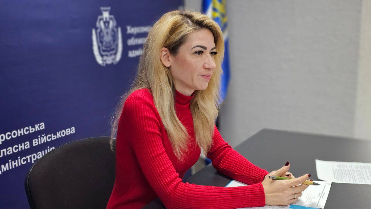 Крупний план жінки зі світлим волоссям, одягненої в червоний гольф, яка сидить за столом під час зустрічі. На задньому плані видно частину синього банера з написом