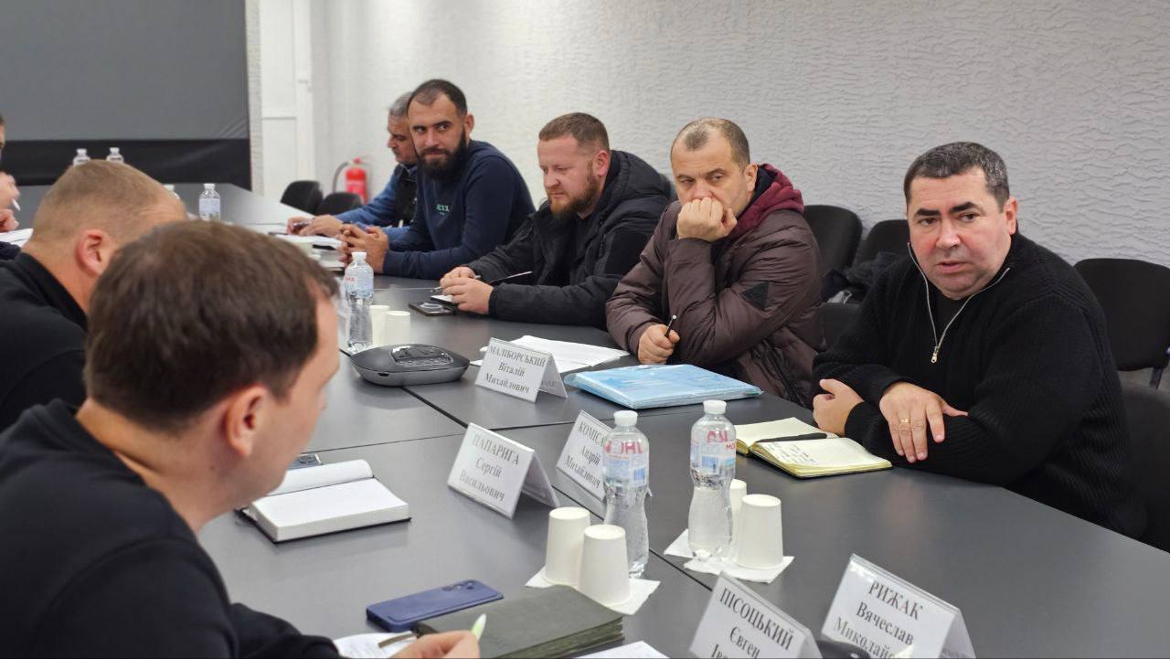 Група чоловіків сидить за столом на нараді, перед ними стоять таблички з прізвищами та іменами. Чоловіки зосереджено слухають або беруть участь в обговоренні.