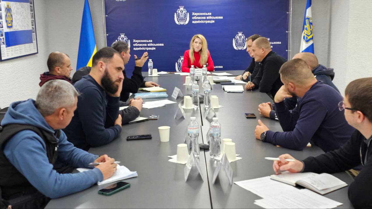 Жінка в червоному светрі сидить на чолі столу, ведучи зустріч із групою чоловіків у неформальному одязі. На задньому плані розташовані прапори України та Херсонської області, а також банер