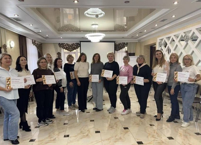 Групова фотографія: чотирнадцять жінок стоять у світлому, елегантно оформленому залі після тренінгу чи навчання, тримаючи в руках сертифікати.