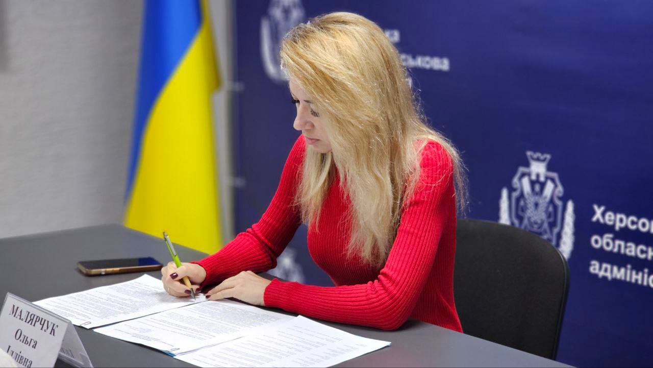 Жінка зі світлим волоссям, одягнена у червоний светр, сидить за столом і зосереджено підписує документи зеленою ручкою. Перед нею на столі лежить мобільний телефон. На синьому фоні позаду неї видно логотип та напис