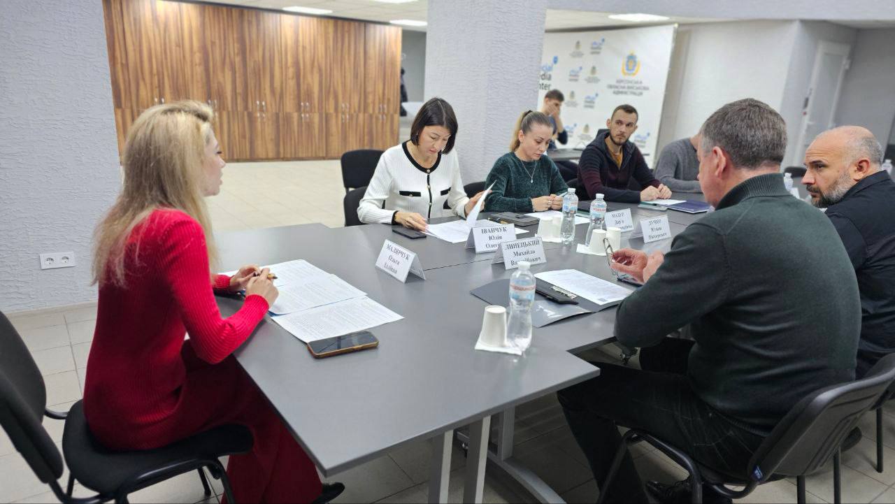 Група з семи людей сидить за довгим сірим столом у приміщенні офісу під час зустрічі. На столі лежать документи та пляшки з водою. Жінка в яскраво-червоному светрі сидить ліворуч, обличчям до чоловіків праворуч. У центрі столу лежать іменні таблички українською мовою.