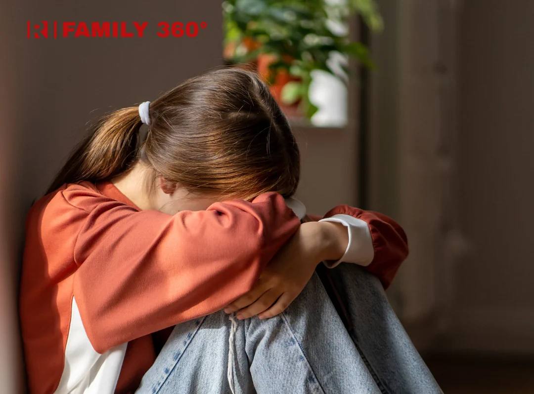 Знімок великим планом, на якому дівчина сидить, обхопивши коліна і сховавши обличчя, притулившись до стіни. На ній помаранчева кофта і джинси. Зображення передає відчуття смутку або самотності.