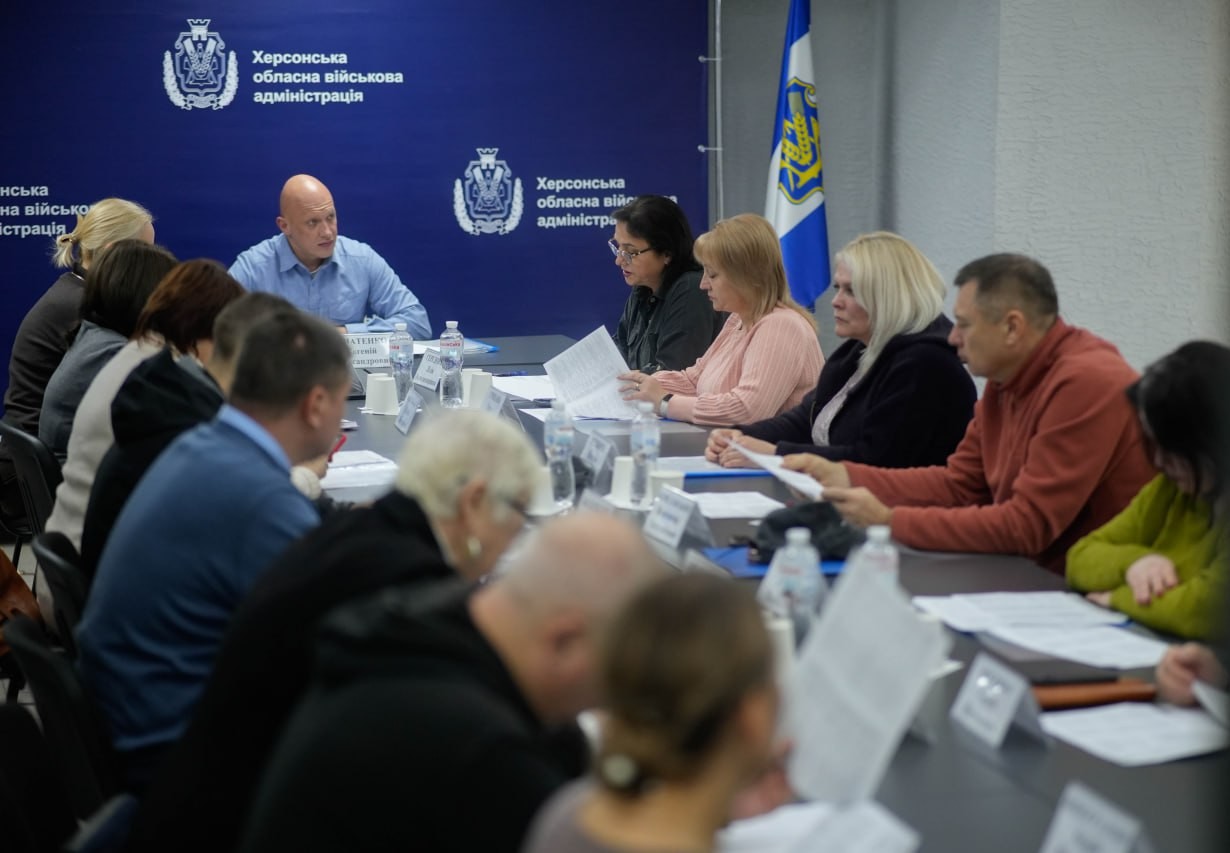 Знімок з наради або засідання за довгим столом. У кадрі багато людей, що сидять навколо столу, тримають папери та слухають чоловіка в блакитній сорочці, який говорить у центрі. На тлі — темно-синій банер з гербом та назвою