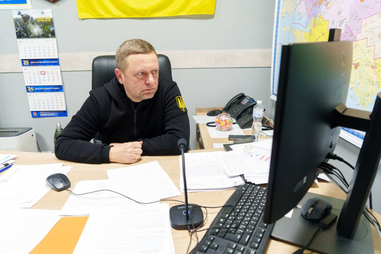 Напівпрофільний знімок чоловіка в чорній кофті з емблемою, який сидить за робочим столом перед монітором комп'ютера. Стіл завалений паперами. На задньому плані видно календар на 2025 рік, державний прапор України та карту. Чоловік, ймовірно, бере участь у відеоконференції або дистанційній нараді, судячи з мікрофона та уважного погляду на екран.