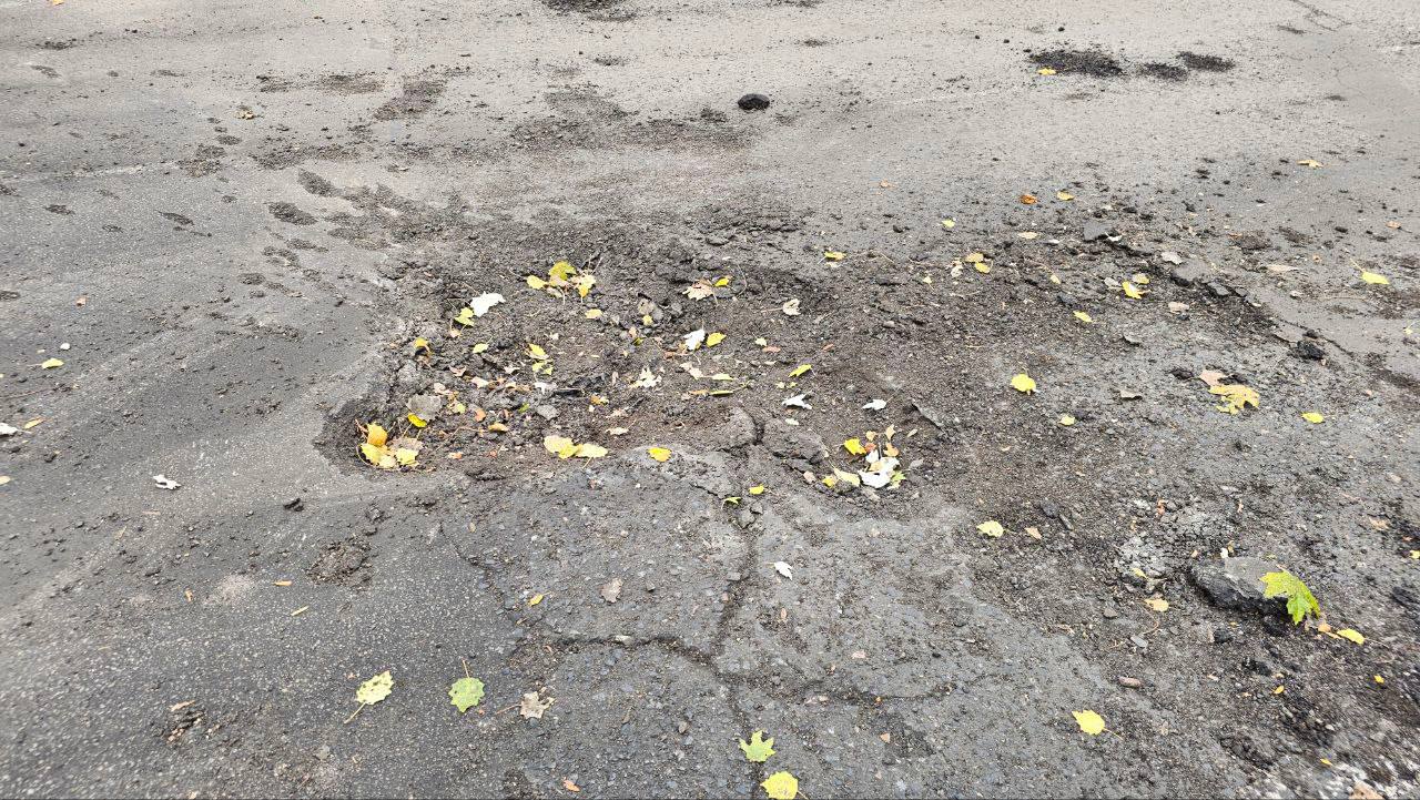 Крупний план значної вибоїни або ділянки пошкодженого асфальтованого дорожнього покриття, наповненої ґрунтом, камінням та опалим жовтим і зеленим листям. Асфальт навколо має тріщини та ознаки руйнування. Знімок зроблено згори.