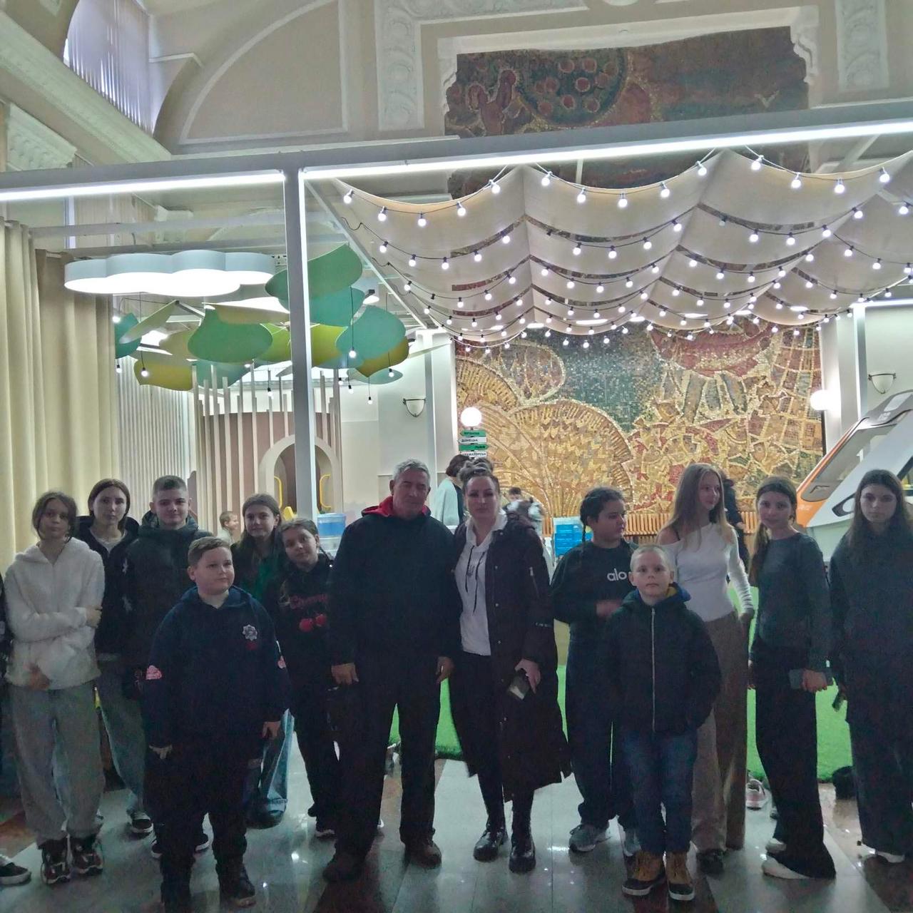 Крупне зображення групи людей, які стоять у приміщенні. На передньому плані: Велика група людей, переважно підлітки та двоє дорослих (чоловік і жінка в центрі), стоять пліч-о-пліч, дивлячись на камеру. Дорослі знаходяться в центрі групи. На задньому плані: Стильно оформлене приміщення з високими стелями. Над головами людей видно інсталяцію, схожу на навіс або ширму з безліччю гірлянд і лампочок, що світяться. За цією інсталяцією знаходиться велике декоративне панно або мозаїка з візерунком у теплих тонах. Ліворуч також видно декоративні підвісні елементи, схожі на хмари.