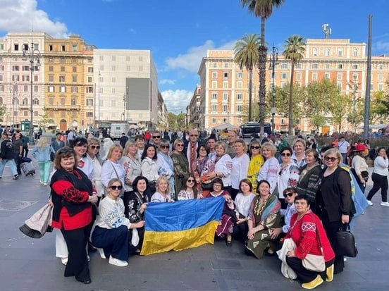 Велика група людей, переважно жінок, стоїть і сидить на міській площі. Більшість жінок одягнені у вишиванки або одяг з елементами української вишивки. Вони тримають великий прапор України (синьо-жовтий) на передньому плані. На задньому плані видно великі європейські будівлі, вулицю, пальму та яскраве небо.