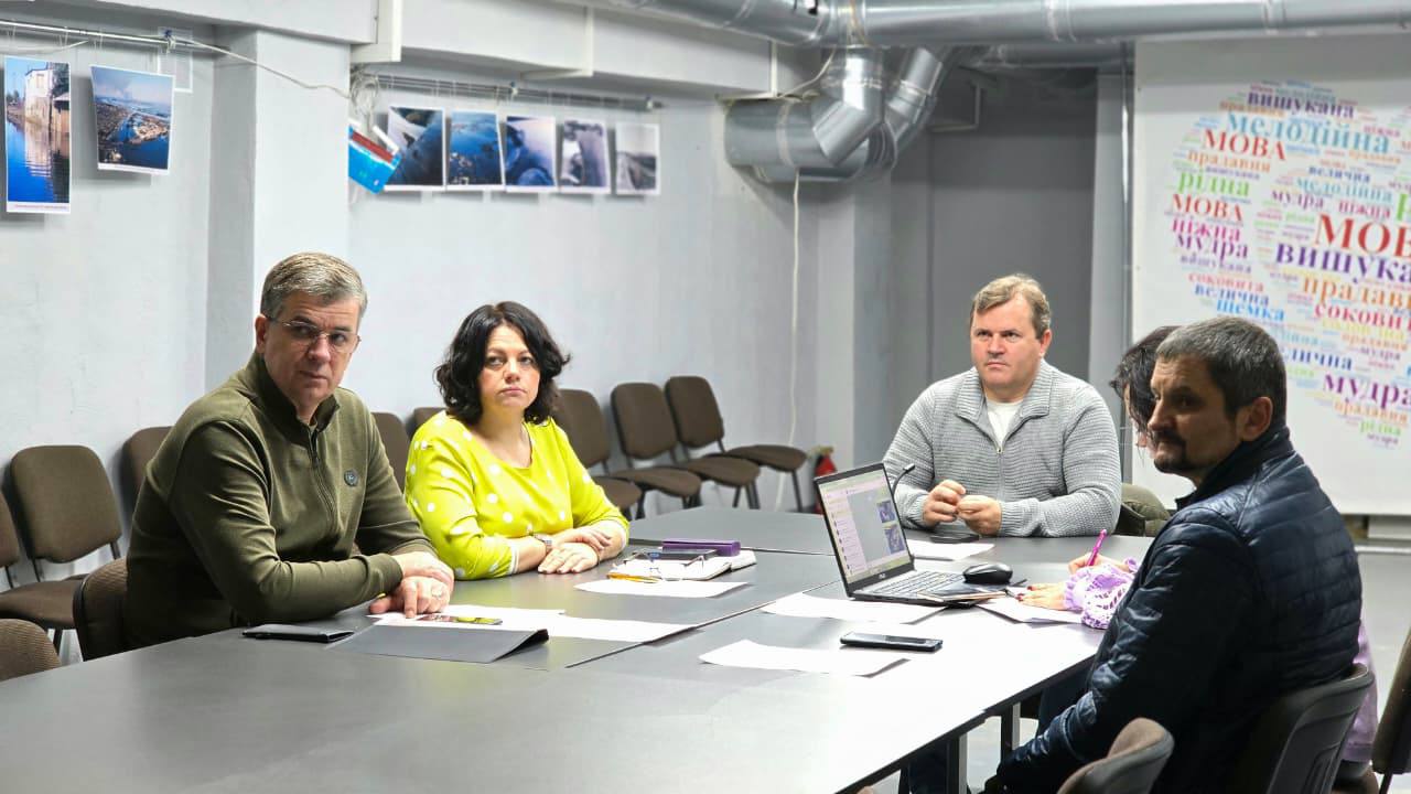 Четверо дорослих (троє чоловіків і одна жінка в яскраво-жовтому светрі) сидять за довгим столом у конференц-залі, беручи участь у зустрічі. На стінах видно фотографії та великий напис-хмара слів з українськими термінами, пов'язаними з мовою.