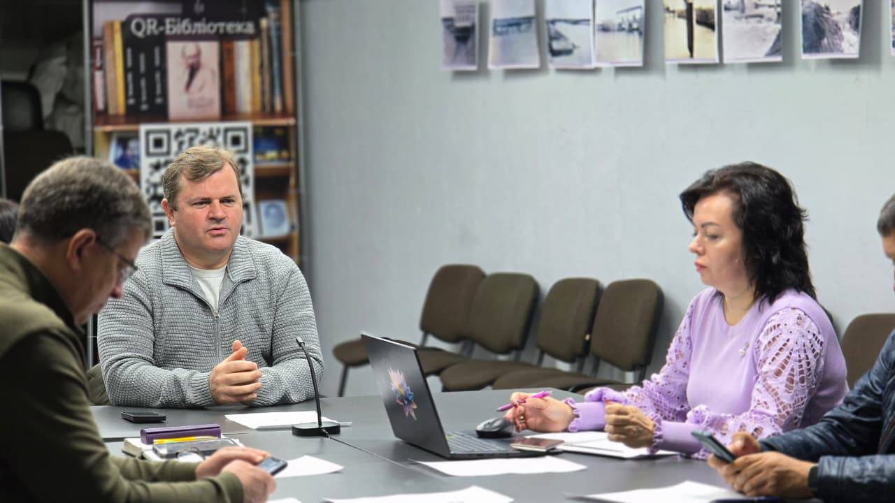 Двоє чоловіків і жінка (у бузковому светрі) сидять за столом під час ділової зустрічі. У центрі уваги чоловік у світло-сірому светрі, що сидить навпроти жінки. На задньому плані видно плакат 