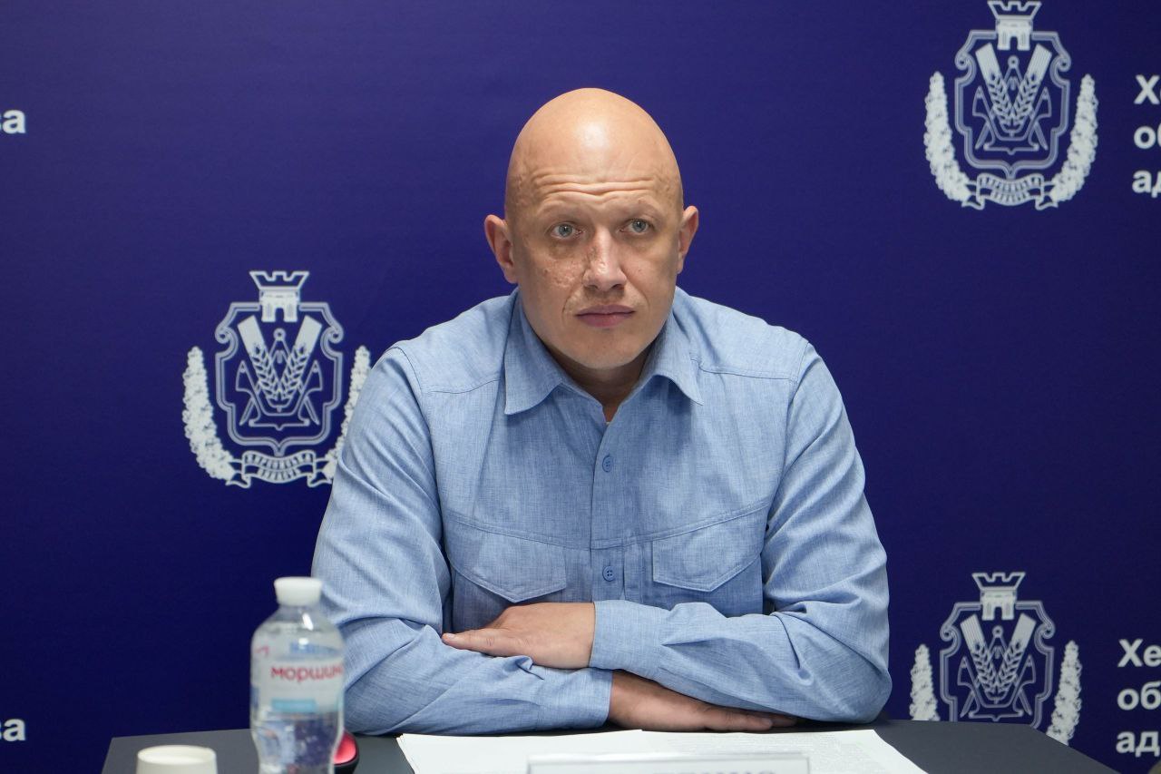 Портретний знімок чоловіка з лисою головою та в блакитній сорочці, який сидить за столом зі схрещеними на ньому руками. Він дивиться прямо в камеру із серйозним виразом обличчя. На задньому плані видно синій пресвол із логотипом та текстом Херсонська обласна військова адміністрація.