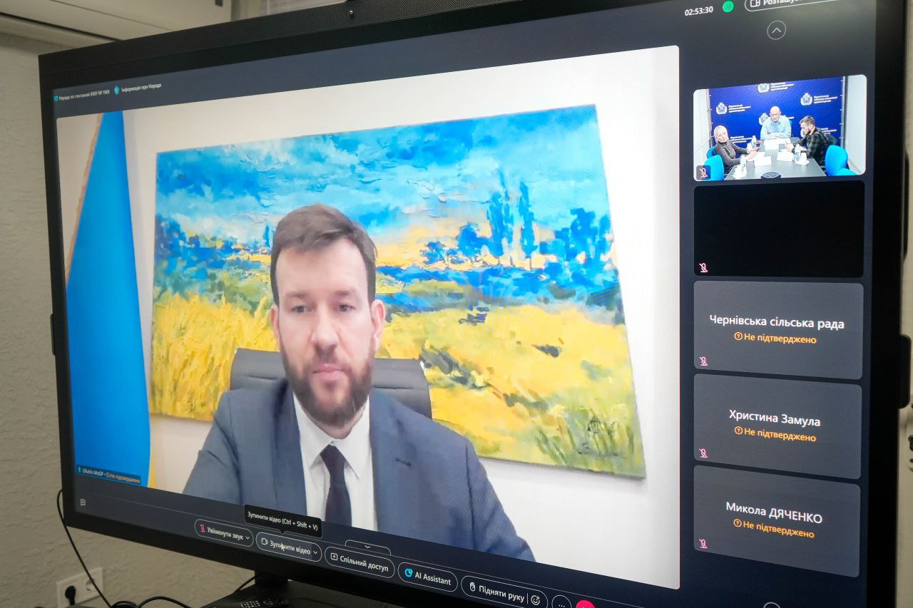 Знімок екрана комп'ютера під час відеоконференції. На головному вікні видно чоловіка в діловому костюмі та краватці, який розмовляє. За ним на стіні висить картина із зображенням жовтого пшеничного поля та синього неба, а також синьо-жовтий прапор України. Праворуч на екрані розташовані вікна інших учасників, зокрема мініатюрне вікно з трьома людьми за столом (зображення, схоже на попередні у цій розмові).