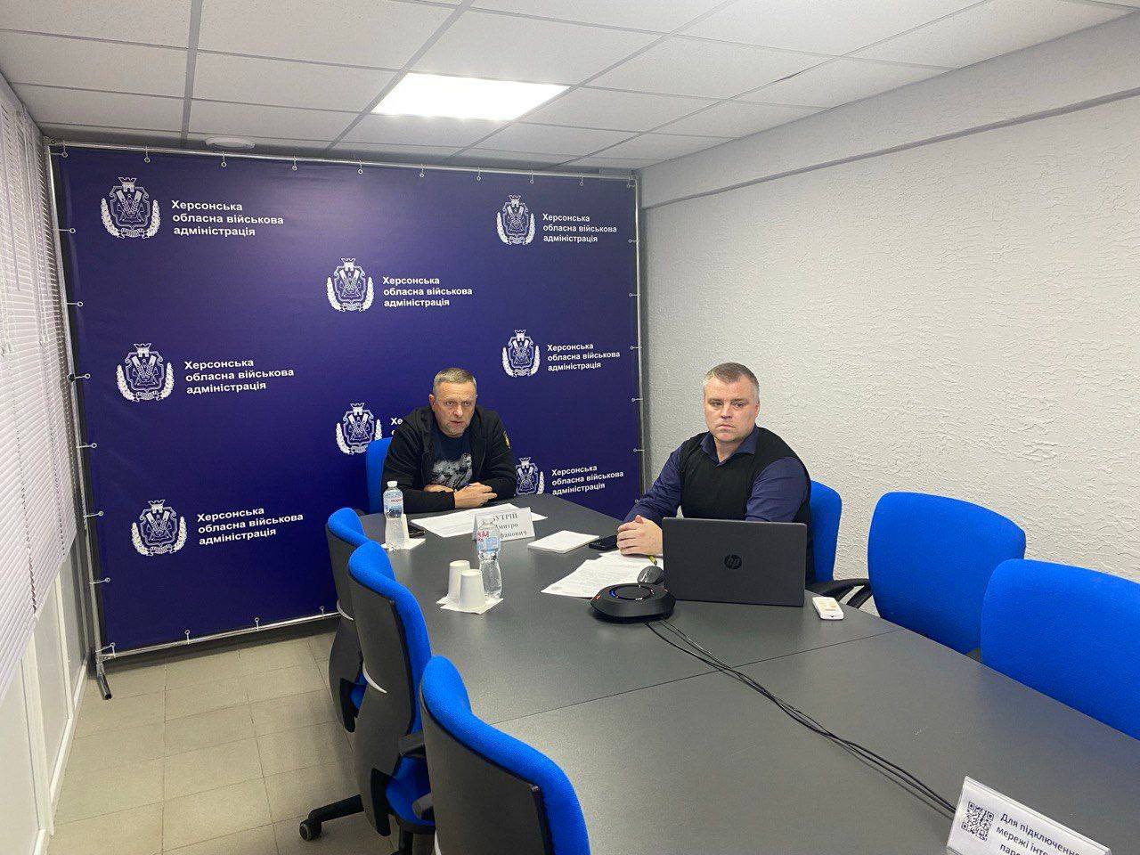 Двоє чоловіків сидять за великим столом для нарад у кімнаті під час онлайн-наради. За ними знаходиться синій пресвол із повторюваним логотипом та написом Херсонська обласна військова адміністрація.