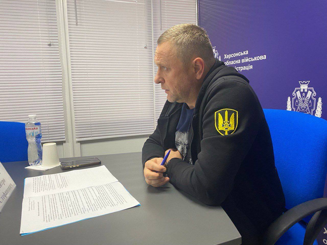 Чоловік середнього віку у чорній кофті з капюшоном та жовто-чорною нашивкою українського герба на рукаві сидить за столом. Він дивиться вбік, тримаючи в руці ручку. Перед ним на столі лежать документи. На задньому плані видно частину синього пресволу з написом Херсонська обласна військова адміністрація.