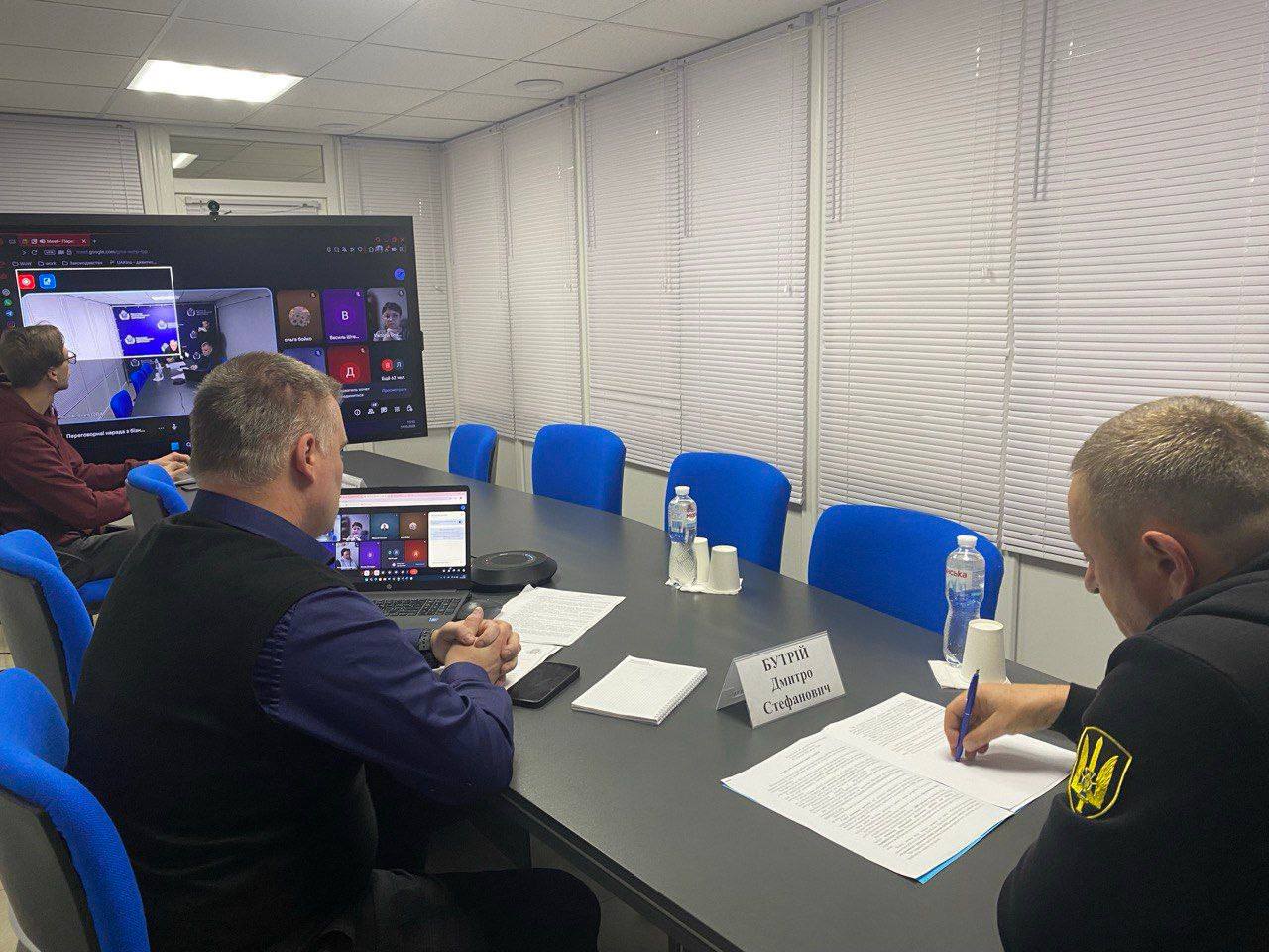 Кадр з робочої наради чи онлайн-конференції. Троє чоловіків сидять за столом, один із них (справа, у чорній кофті з гербом на рукаві) підписує документ. Інший чоловік (ліворуч, спиною) використовує ноутбук. На великому екрані на стіні видно учасників онлайн-зустрічі.