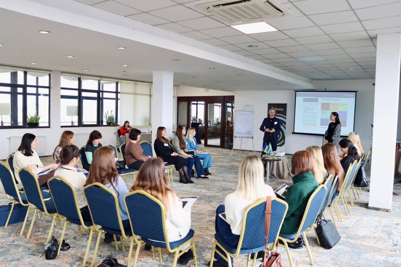 Група людей, переважно жінок, сидить у півколі на стільцях із синьою оббивкою у просторій, світлій конференц-залі з великими вікнами. Перед ними стоять двоє доповідачів — чоловік та жінка, які презентують інформацію на екрані з проектора. На трибуні перед чоловіком видно невеликий прапор України.