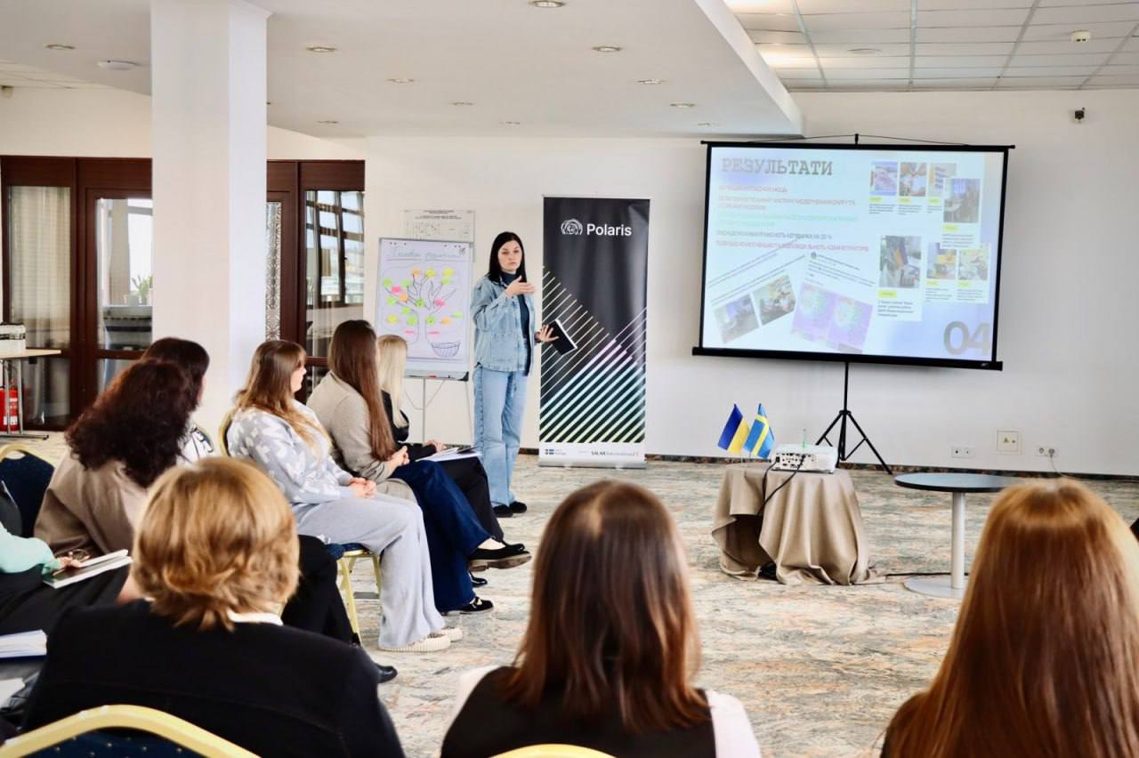 Жінка в джинсовому одязі стоїть перед групою слухачок, проводячи презентацію у світлому конференц-залі. Вона жестикулює, стоячи поруч із рекламним банером «Polaris». На проекційному екрані відображаються «РЕЗУЛЬТАТИ» з текстом та зображеннями. На невеликому столику праворуч видно два мініатюрні прапори України та Швеції.