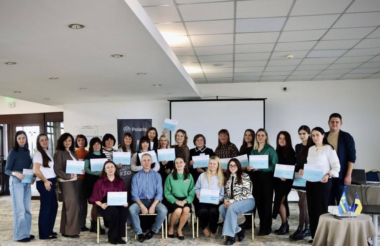 Група з приблизно 25 людей, більшість з яких жінки, позує для спільної фотографії у приміщенні конференц-залу. Більшість із них тримають у руках сертифікати або документи. За ними видно банер «Polaris», проекційний екран та столик із прапорами України та Швеції.
