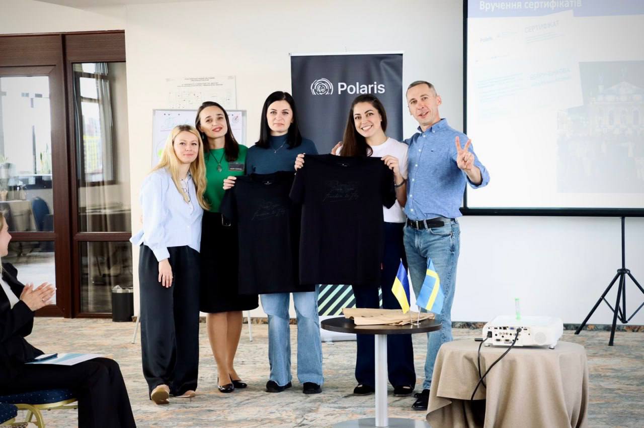 Четверо жінок та один чоловік стоять разом, посміхаючись, після події у конференц-залі. Дві жінки в центрі тримають чорні футболки з принтом. На задньому плані видно банер «Polaris» та проекційний екран з написом «Вручення сертифікатів». Поруч із ними на невеликому столику стоять два мініатюрні прапори України та Швеції.