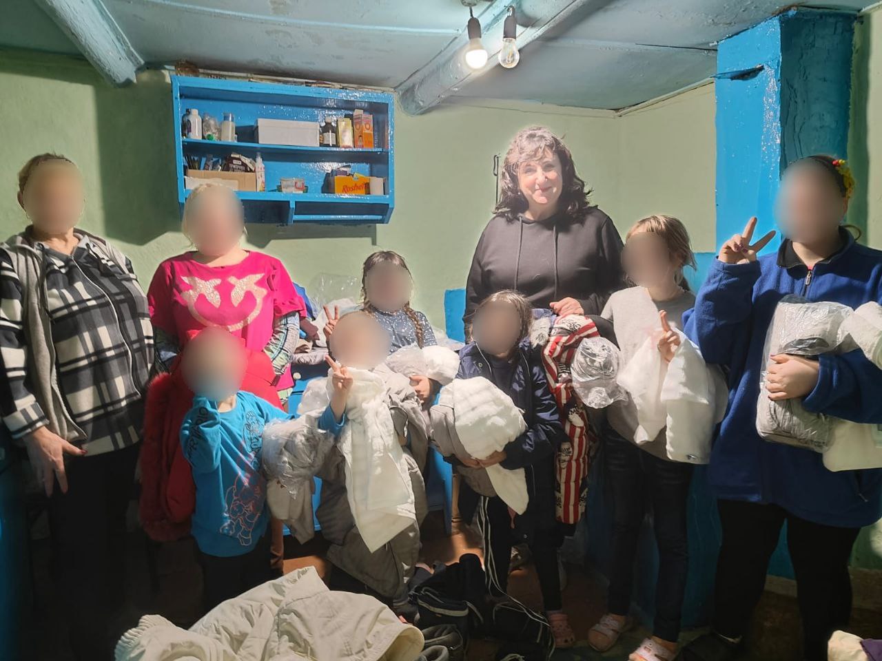 Група жінок і дітей у приміщенні отримує допомогу у вигляді одягу та речей. Діти тримають білі згортки, а дорослі стоять поруч, посміхаючись.