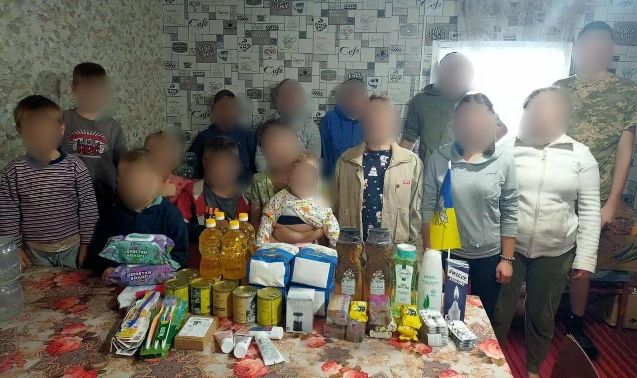 Групове фото, на якому близько десятка дорослих та дітей стоять за столом із зібраними предметами гуманітарної допомоги, включаючи консерви, олію, вологі серветки, засоби гігієни та мийні засоби.