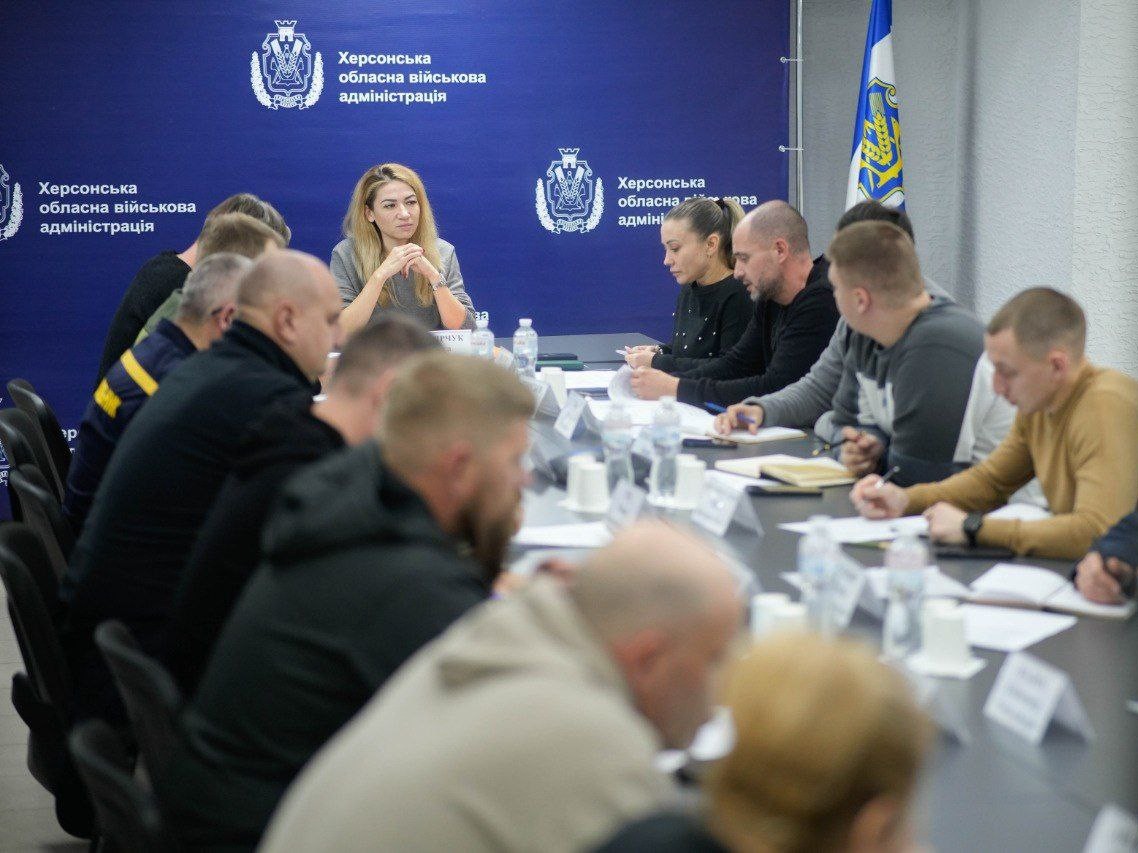 Група людей сидить за великим столом для нарад у кімнаті, ймовірно, під час зустрічі або брифінгу. У центрі уваги — жінка з довгим світлим волоссям, яка сидить за столом, склавши руки. Більшість учасників — чоловіки, які сидять спиною або боком до камери. На задньому плані видно синій пресвол Херсонська обласна військова адміністрація та синьо-білий прапор.