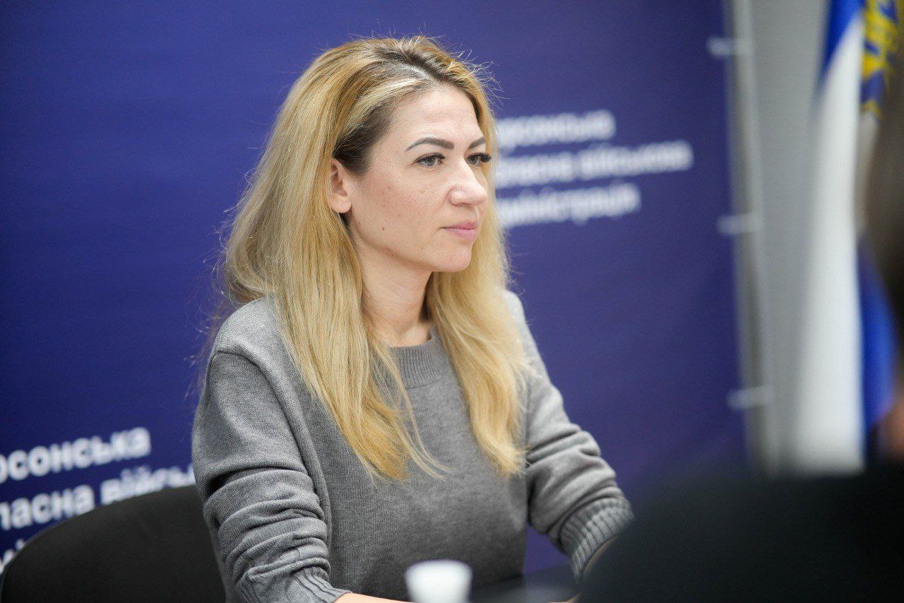 Портретний знімок жінки з довгим світлим волоссям, яка сидить за столом. Вона одягнена в сірий светр і дивиться вправо. На задньому плані видно синій пресвол із написом, що стосується Херсонської обласної військової адміністрації.