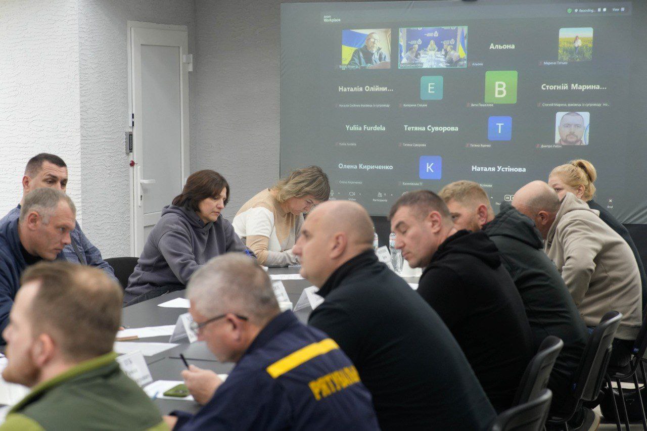 Група людей сидить за довгим столом для нарад, уважно слухаючи або працюючи з документами. На задньому плані видно великий екран, де відбувається відеоконференція з кількома учасниками.