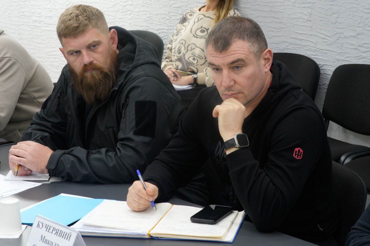 Двоє чоловіків сидять за столом під час наради. Чоловік праворуч у чорній кофті з шевроном-тризубом робить нотатки в блокноті. Чоловік ліворуч має густу руду бороду і також одягнений у чорне.