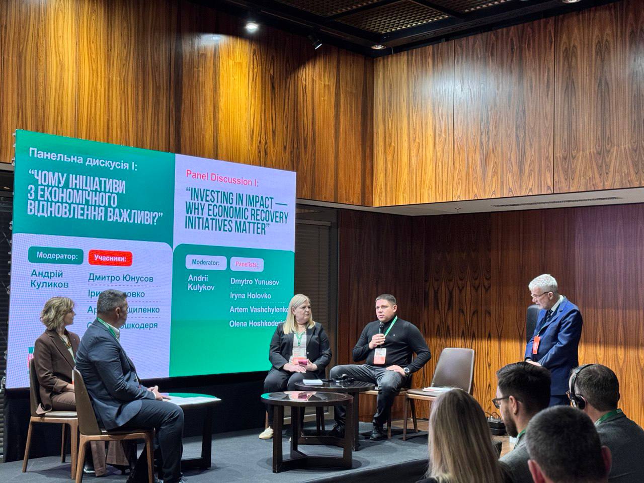 Панельна дискусія на тему: ЧОМУ ІНІЦІАТИВИ З ЕКОНОМІЧНОГО ВІДНОВЛЕННЯ ВАЖЛИВІ? Четверо учасників сидять на сцені перед екранами зі списком модератора та спікерів. П'ятий учасник, старший чоловік у костюмі, стоїть праворуч, розмовляючи з ними. Видно частину глядачів на передньому плані.