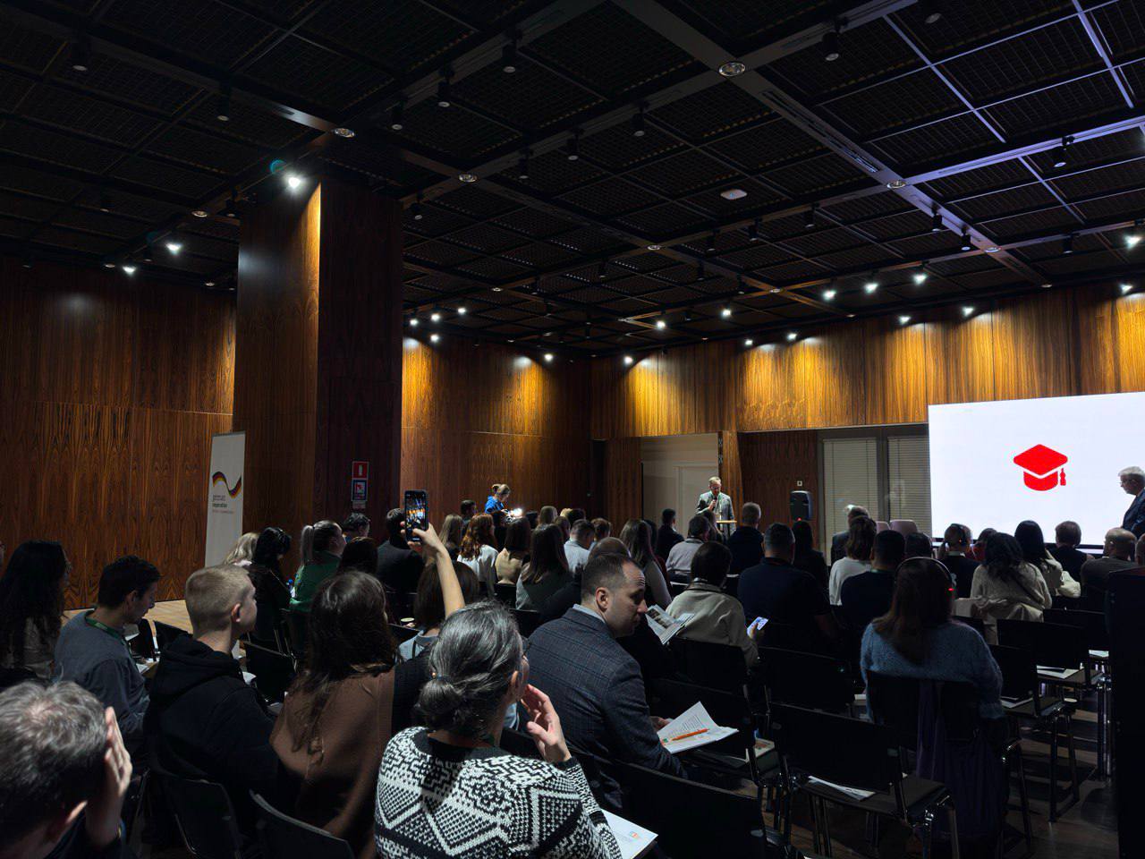 Вигляд на великий, напівтемний конференц-зал із дерев'яними стінами та стелею. Численні слухачі сидять рядами, звернені до сцени. На сцені стоїть доповідач, а на великому екрані праворуч відображено червону академічну шапочку (конфедератку), що символізує освіту чи випуск.