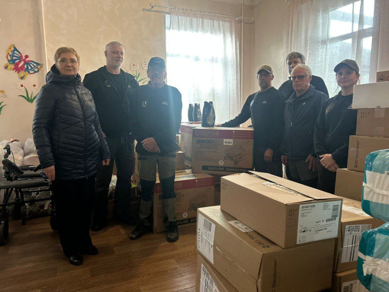 Група з восьми людей (двоє жінок та шестеро чоловіків), переважно в темному одязі та бейсболках, стоять у приміщенні поруч із великою кількістю гуманітарної допомоги. Допомога включає картонні коробки та коробки з генераторами. Люди стоять у кімнаті зі світлими стінами та великим вікном із тюлевими шторами на задньому плані.