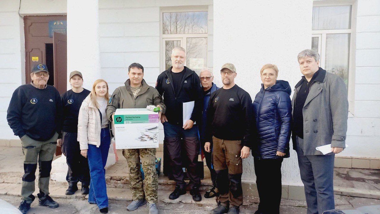 Група з восьми людей, включно з військовим у камуфляжі, стоять перед білою будівлею, ймовірно, для передачі гуманітарної допомоги. Військовий у центрі тримає коробку з принтером HP. Учасники групи вдягнені у різний одяг, дехто має однакові темні футболки та бейсболки з емблемами, що вказує на приналежність до однієї організації.