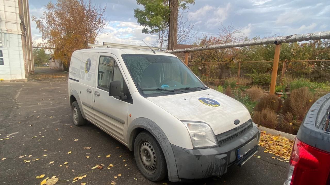 Білий фургон Ford Transit Connect припаркований на асфальтованій ділянці з опалим осіннім листям. На капоті фургона видно емблему або маркування, і на ньому встановлений багажник на даху. Фургон виглядає брудним і робочим. На задньому плані — паркан, дерева та похмуре небо.