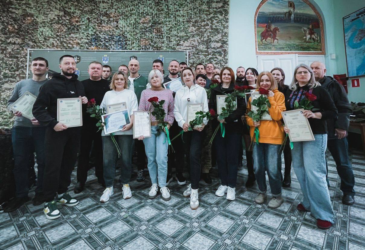 Велика група чоловіків і жінок стоять разом у приміщенні на тлі стіни з камуфляжним малюнком. Усі тримають подяки або сертифікати, а жінки додатково тримають по букету червоних троянд.