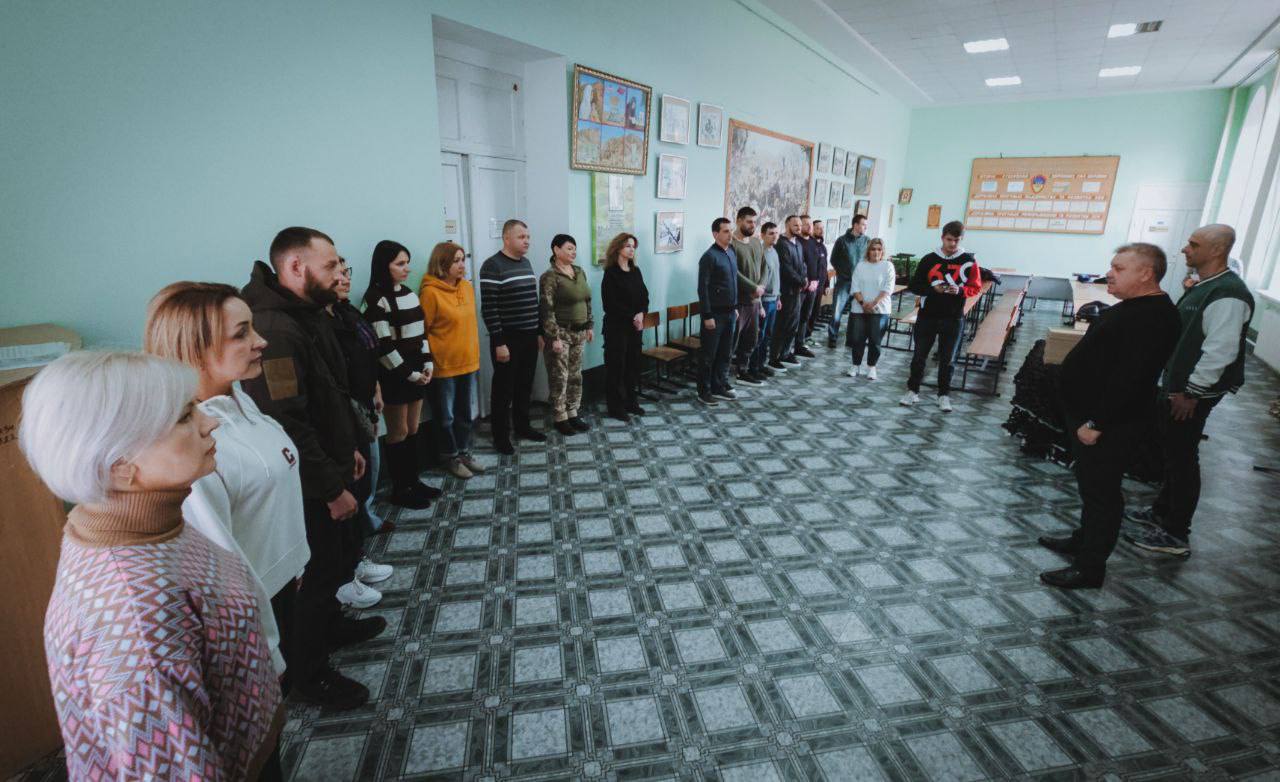 Група з приблизно 20 людей стоїть рівним строєм у світлій навчальній кімнаті з бірюзовими стінами та підлогою з геометричним візерунком. Вони звернені до двох чоловіків, що стоять праворуч, які, ймовірно, проводять інструктаж або церемонію. На стінах видно картини, плакати та стенди.