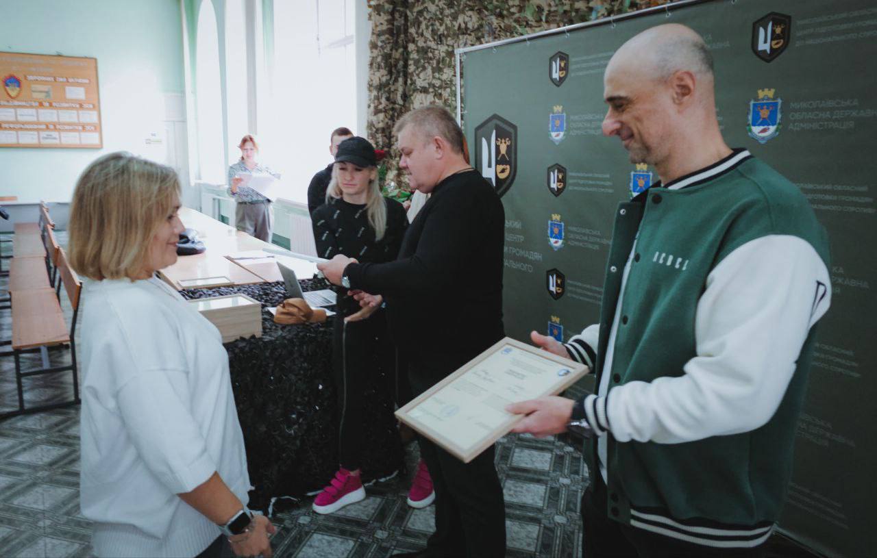 Церемонія вручення нагород або документів у приміщенні. Чоловік у зелено-білій куртці-бомбері вручає сертифікат жінці у білому светрі. Поруч стоять ще двоє людей (чоловік у чорному светрі та жінка у кепці), які також беруть участь у процесі.