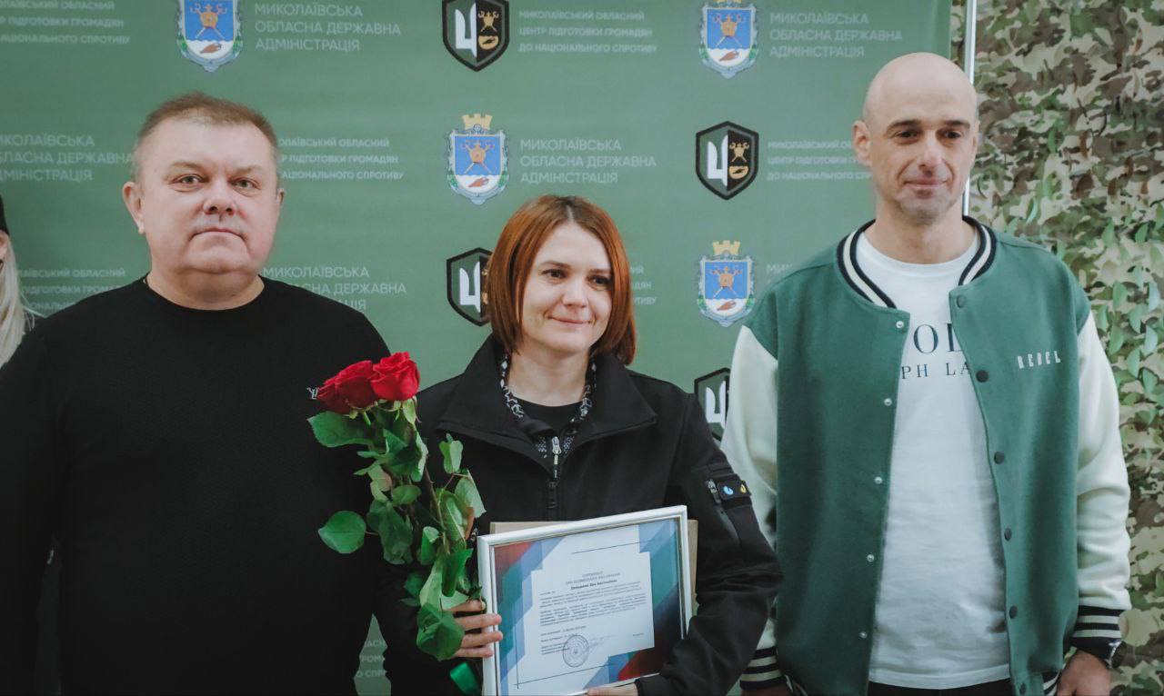 Троє людей — двоє чоловіків та одна жінка — стоять разом для фотографії. Жінка в центрі тримає в руках сертифікат або нагороду та букет червоних троянд, посміхаючись. Чоловіки стоять по обидва боки від неї.