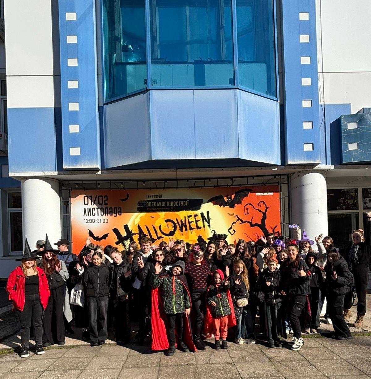 Велика група підлітків та молоді в костюмах та атрибутах на честь свята Хелловін стоїть перед будівлею з великим рекламним банером p великим шрифтом HALLOWEEN на помаранчево-чорному тлі з силуетами кажанів та дерев. Більшість людей махають руками.