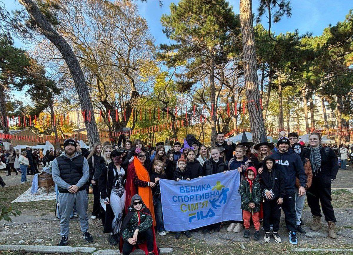 Велика група молодих людей у карнавальних та спортивних костюмах тримає банер із написом ВЕЛИКА СПОРТИВНА СІМ'Я FILA на тлі осіннього парку з високими деревами.