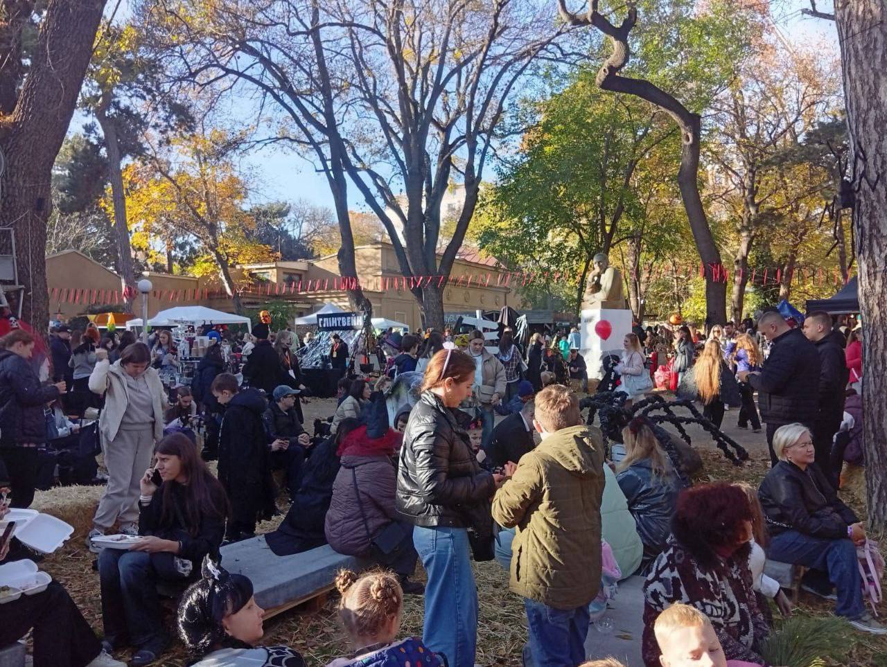 Сцена осіннього фестивалю або ярмарку на відкритому повітрі в парковій зоні з великою кількістю відвідувачів. Люди сидять на сіні та лавках, спілкуються та їдять. На задньому плані видно торгові намети, осінні дерева з жовтим листям і, ймовірно, пам'ятник або скульптуру. Серед натовпу можна помітити елементи святкування Хелловіну (наприклад, роги або прикраси).