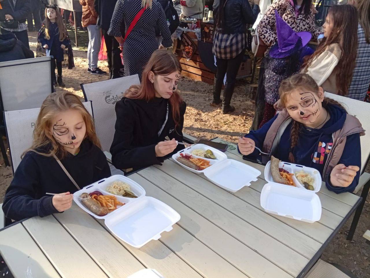 Крупний план трьох дівчат-підлітків у гримі, що імітує черепи на Геловін, які сидять за столом на вулиці та їдять з одноразових контейнерів.