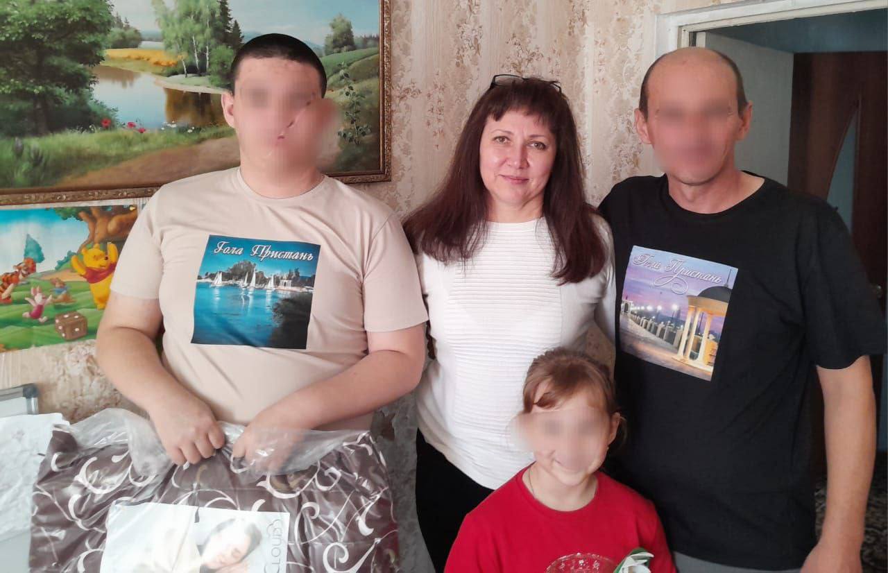 Сімейний портрет у приміщенні: мати у білому светрі, батько і син (обличчя обох розмиті) у футболках з написом Тиха Пристань, та маленька дівчинка (обличчя розмите) у червоній футболці, що стоїть попереду. Син тримає в руках предмет, схожий на ковдру чи постільну білизну в прозорій упаковці. На стіні позаду висить картина з пейзажем.