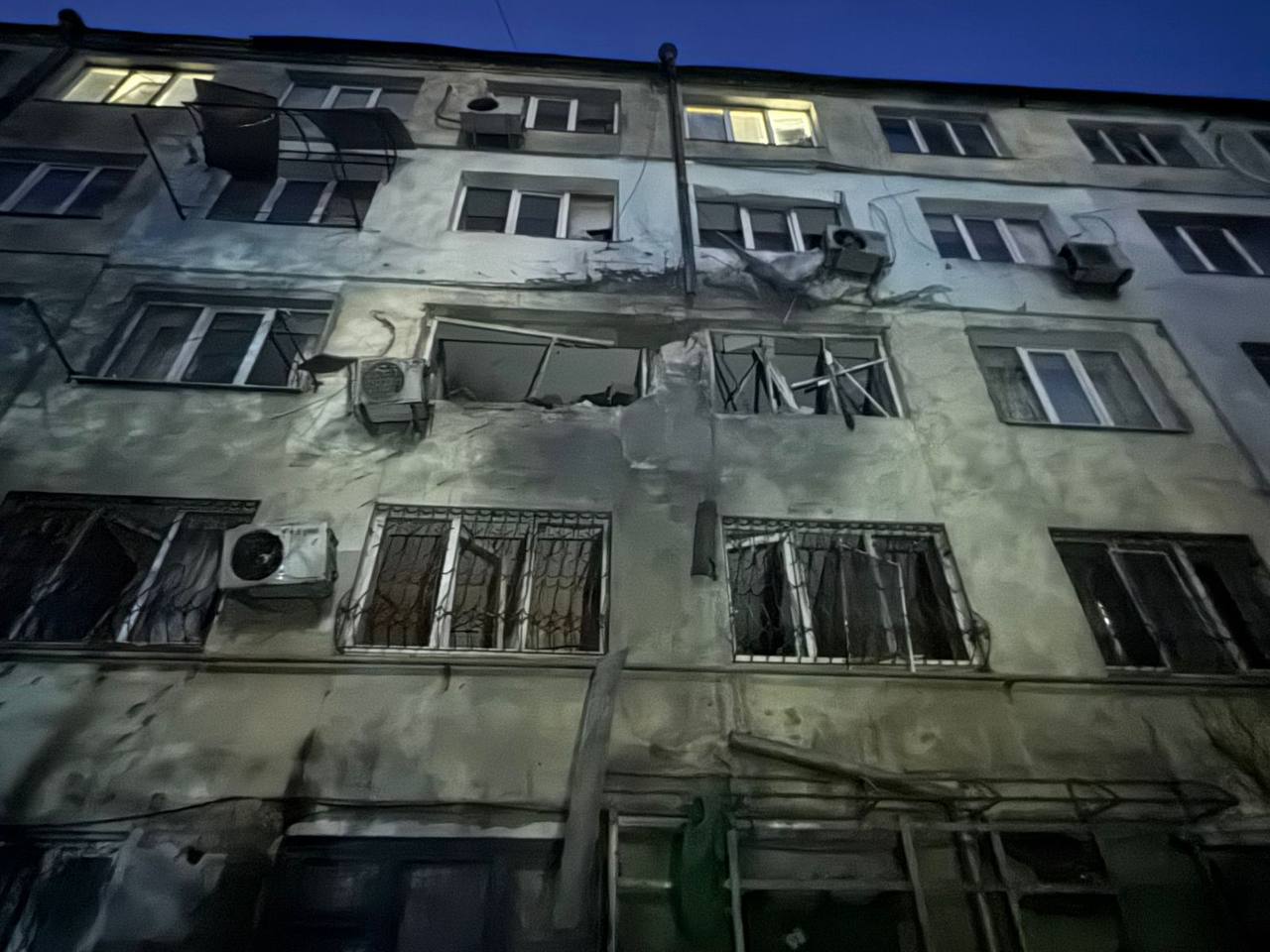 Нічний знімок багатоквартирного житлового будинку зі слідами пошкоджень, ймовірно, внаслідок обстрілу. Видно вибиті вікна, розбиті рами, пошкоджені кондиціонери та сліди кіптяви на фасаді навколо центральної частини будівлі, що вказує на пряме влучання або вибух. У деяких вікнах горить світло.